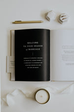 Load image into Gallery viewer, Open Marriage Journal with a black page displaying text on a white surface with a candle and pen.
