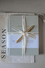 Load image into Gallery viewer, Sage green Season Journals Pregnancy Journal wrapped in a cream ribbon, tied with a gold pen, on top of a fabric dust bag.