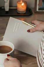 Load image into Gallery viewer, Woman holding Everyday Positive Journal, gold pen, cup of coffee with candle in the background
