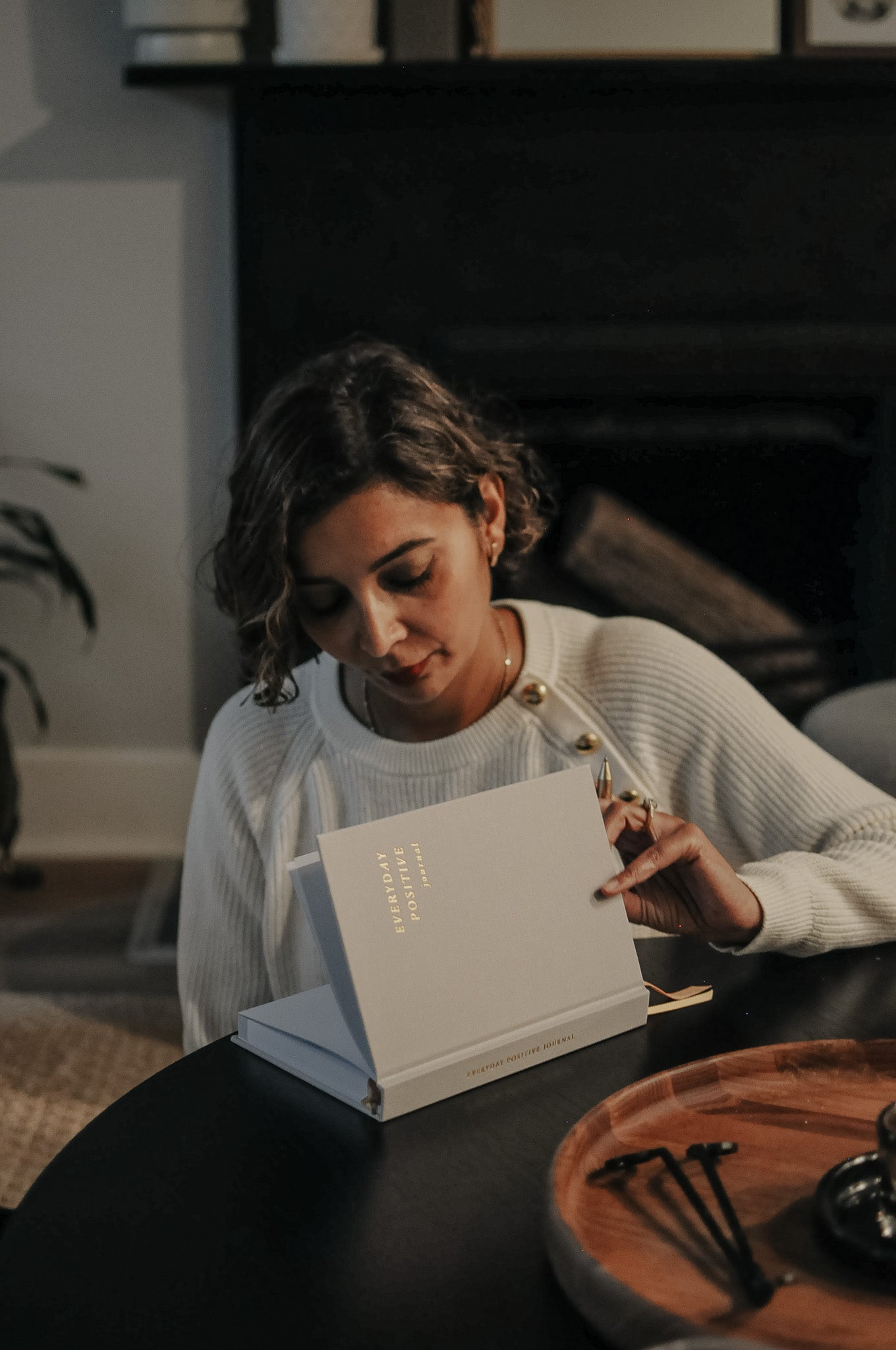 Woman opening the Everyday Positive Journal in a cozy room with a fireplace.