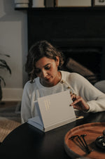 Load image into Gallery viewer, Woman opening the Everyday Positive Journal in a cozy room with a fireplace.
